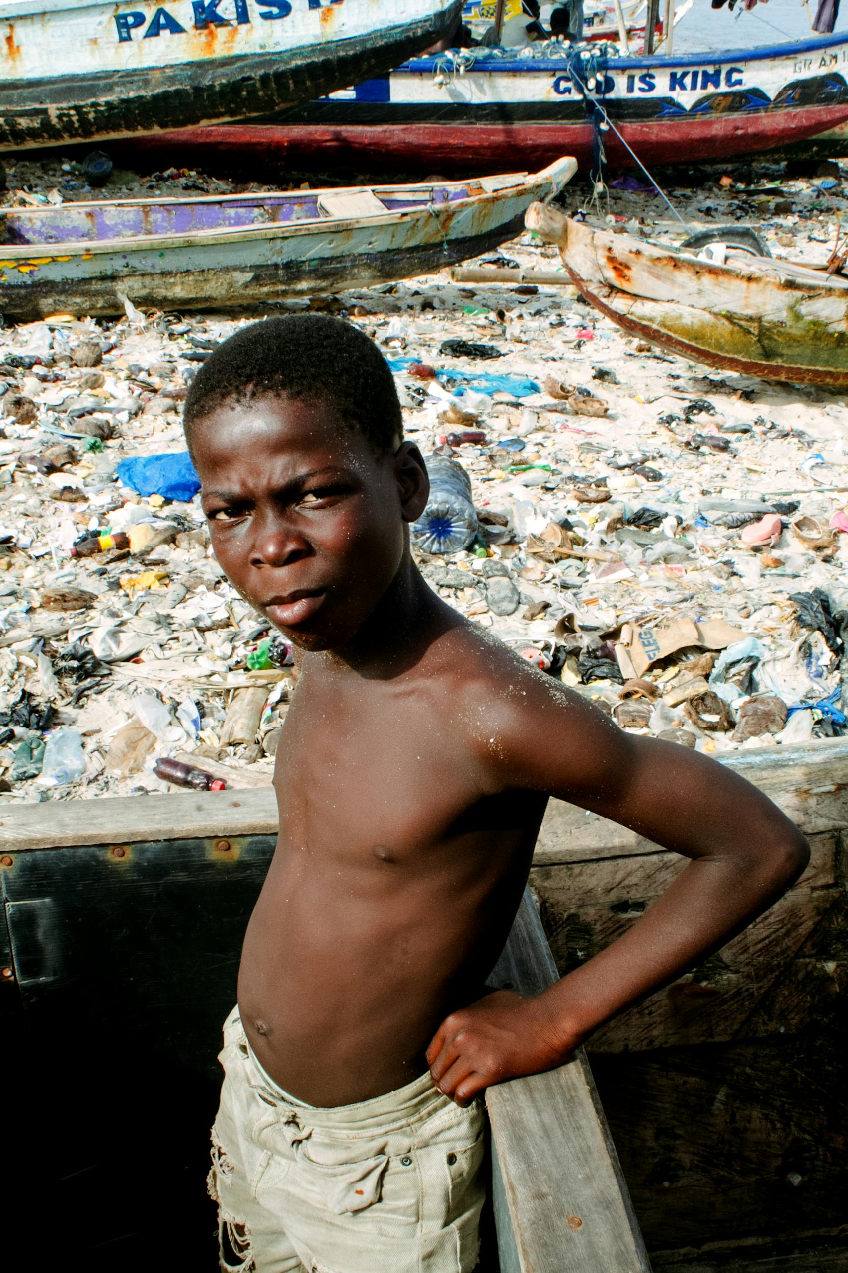 A young boy stands by a polluted river filled with trash and boats under the sunlight.